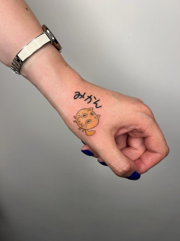 Kolorowy, przymrużający oko tatuaż w stylu kawaii – napis „みかん" (mikan – mandarynka) i uroczy pomarańczowy kotek