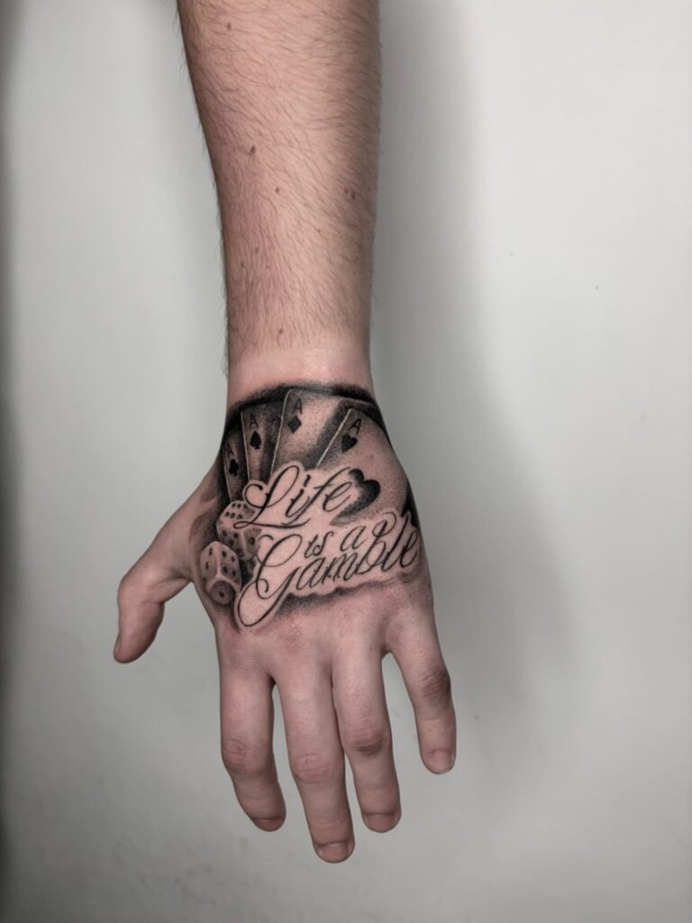 Napis „Life is a Gamble” z asami i kostką do gry – mocna, czytelna kompozycja typograficzna na nadgarstku